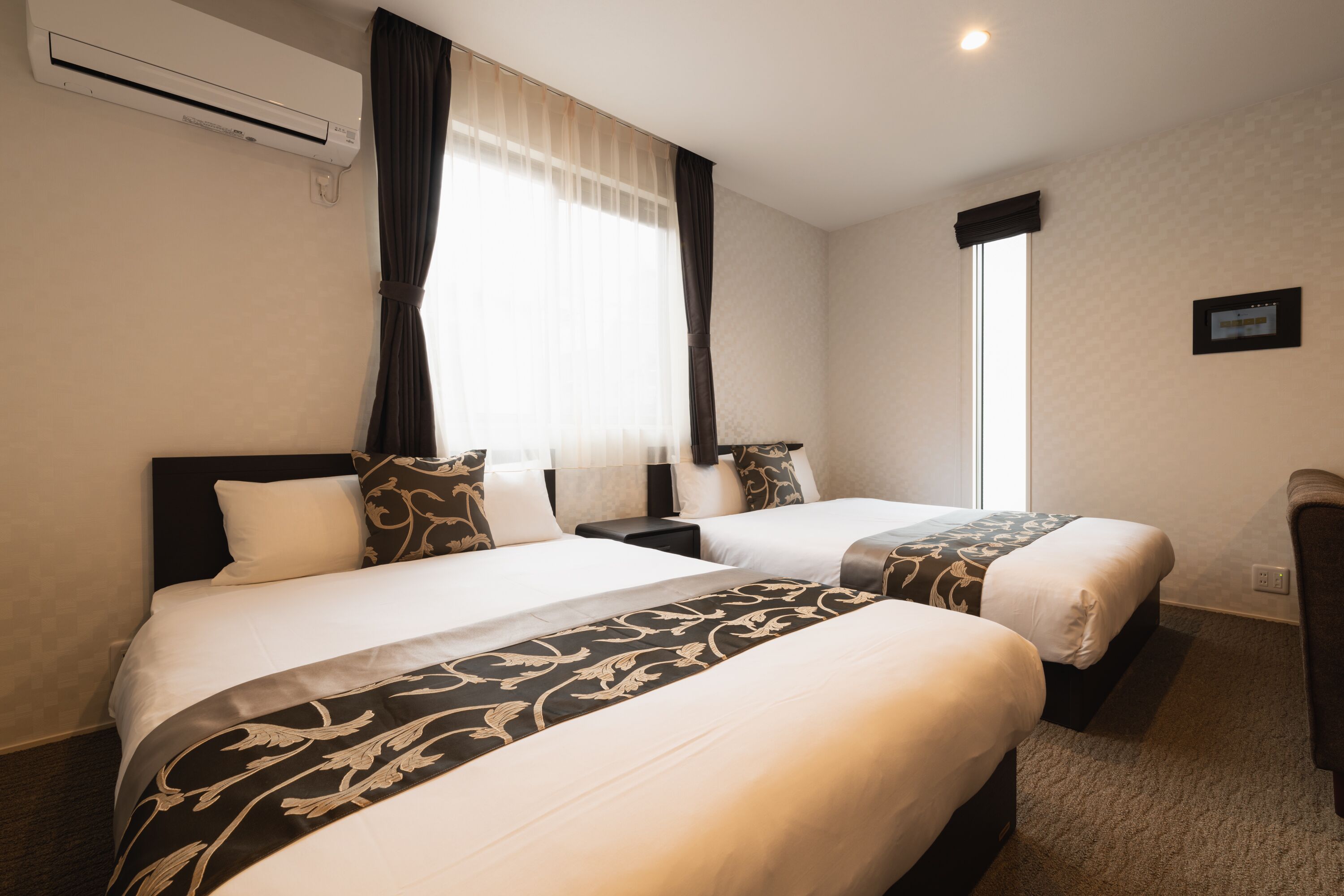 Une chambre d’hôtel avec deux lits, une grande fenêtre avec des rideaux, un climatiseur et un petit bureau avec une chaise.