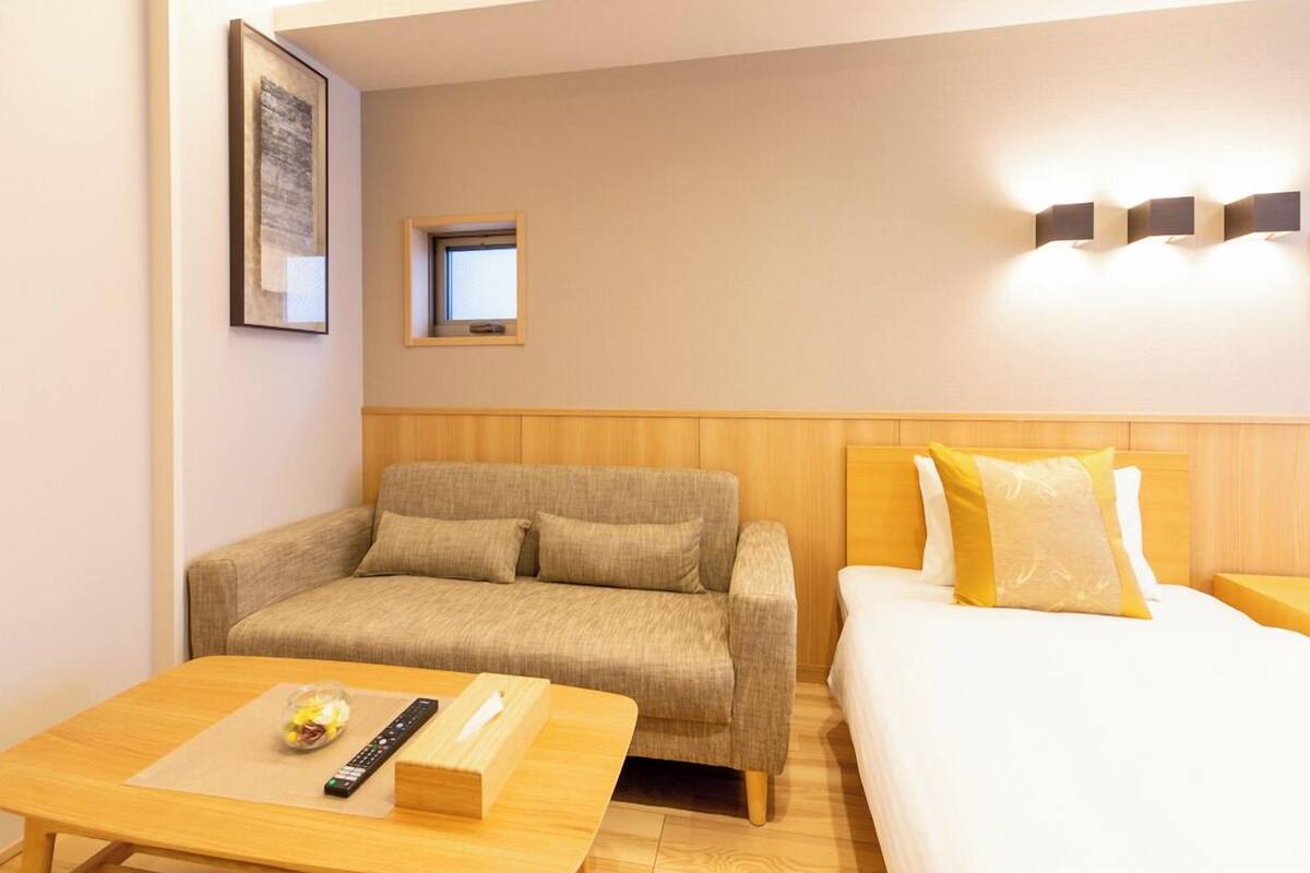 A hotel room with a sofa, a bed, a small table with a remote control, and a picture on the wall.