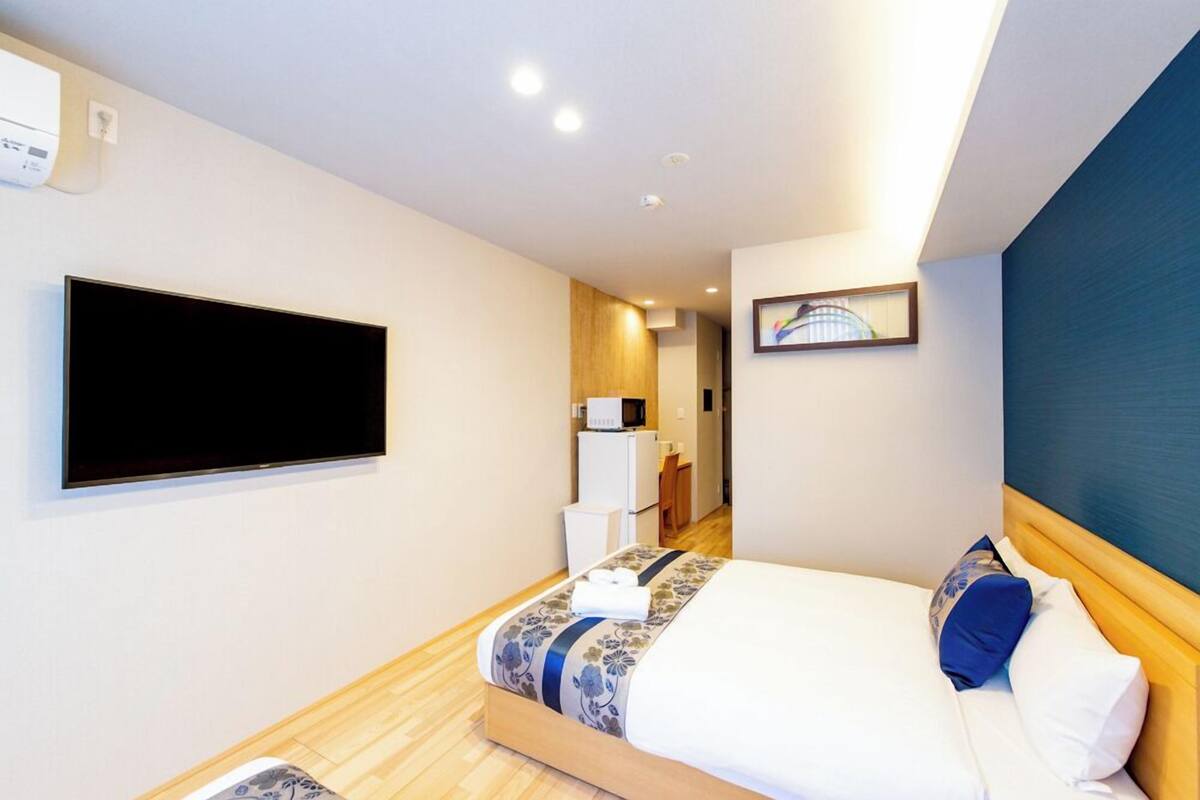A hotel room with a bed, a flat-screen TV, a desk with a computer, and a small kitchenette.