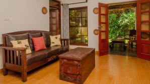 Uma sala de estar com um sofá de madeira, uma mesa pequena de madeira e vista para um jardim através de uma porta aberta.