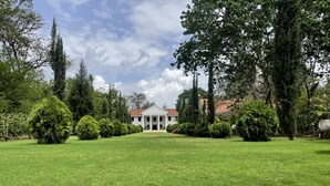 Um edifício grande e branco com fachada simétrica e entrada central, rodeado por jardins bem cuidados e diversas árvores.