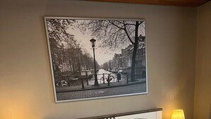 Una cama doble con cabecera, dos mesitas de noche con lámparas, un cuadro en la pared que muestra un paisaje urbano y suelo de madera.