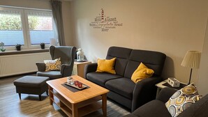 Una sala moderna con un sofá gris oscuro, dos cojines amarillos, una mesa de centro de madera y un sillón gris. Contra la pared, una pequeña mesa con una lámpara blanca y un objeto decorativo.