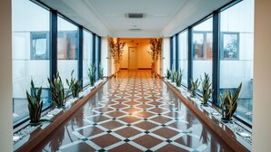 A long, polished hallway with a checkered floor, flanked by large windows and potted plants.