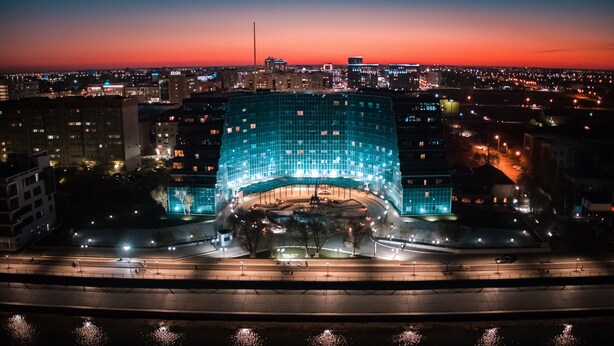 A modern building with a curved glass facade, surrounded by a well-lit plaza with benches and a fountain.