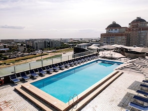 Une piscine sur le toit, dotée de chaises longues, offrant une vue sur le paysage urbain, et un grand bâtiment à l’architecture caractérisée par des dômes en arrière-plan.