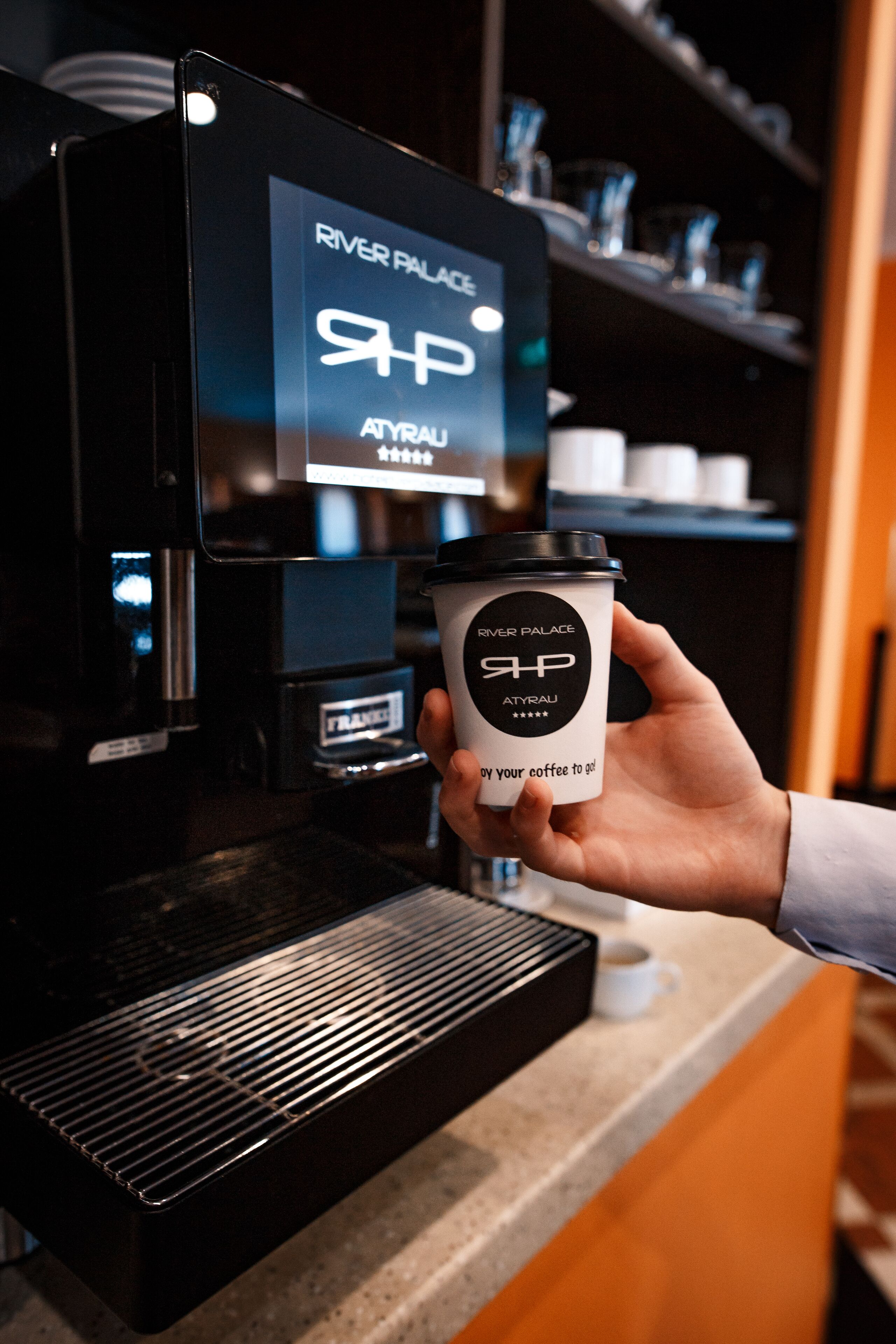A hand holding a coffee cup with the logo "River Palace" and "Atyralu" on a coffee machine.