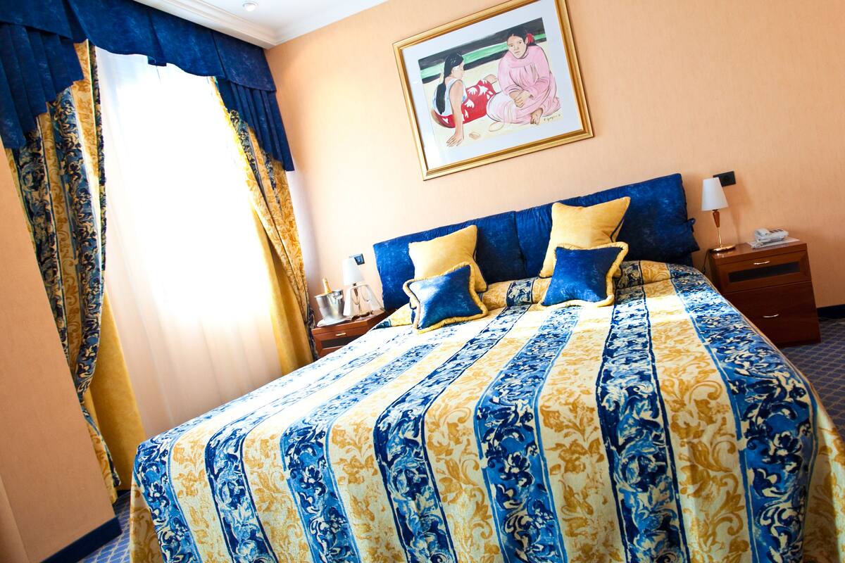Quarto de hotel com uma cama grande, mesa de cabeceira, abajur e um quadro na parede.