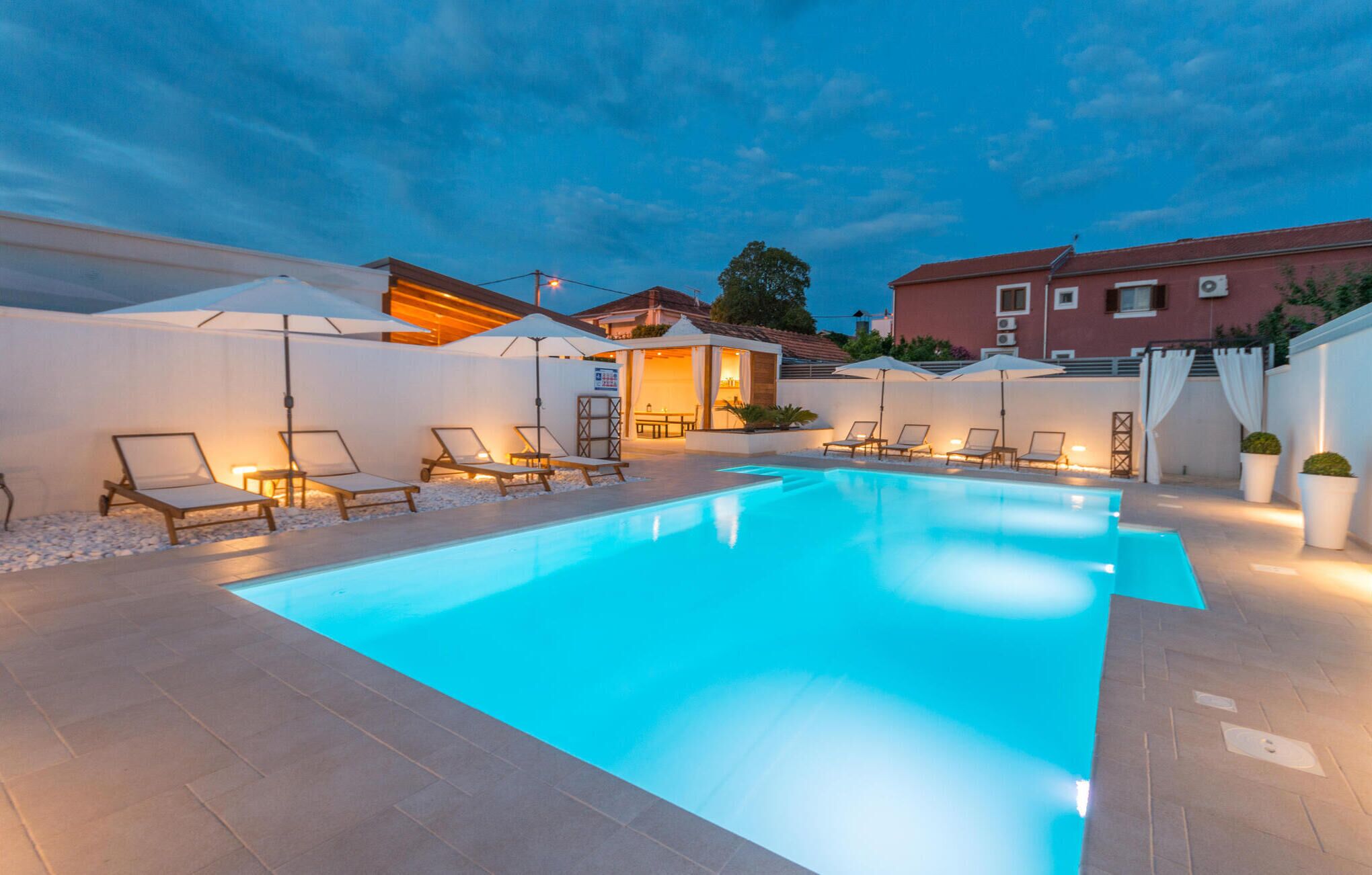 Une piscine entourée de chaises longues et de parasols, le tout bordé d’un mur blanc et avec un bâtiment visible au loin.