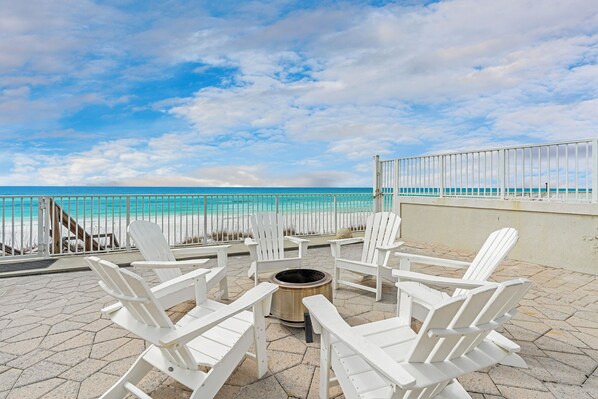 A patio with white Adirondack chairs and a fire pit, overlooking a beach and ocean.