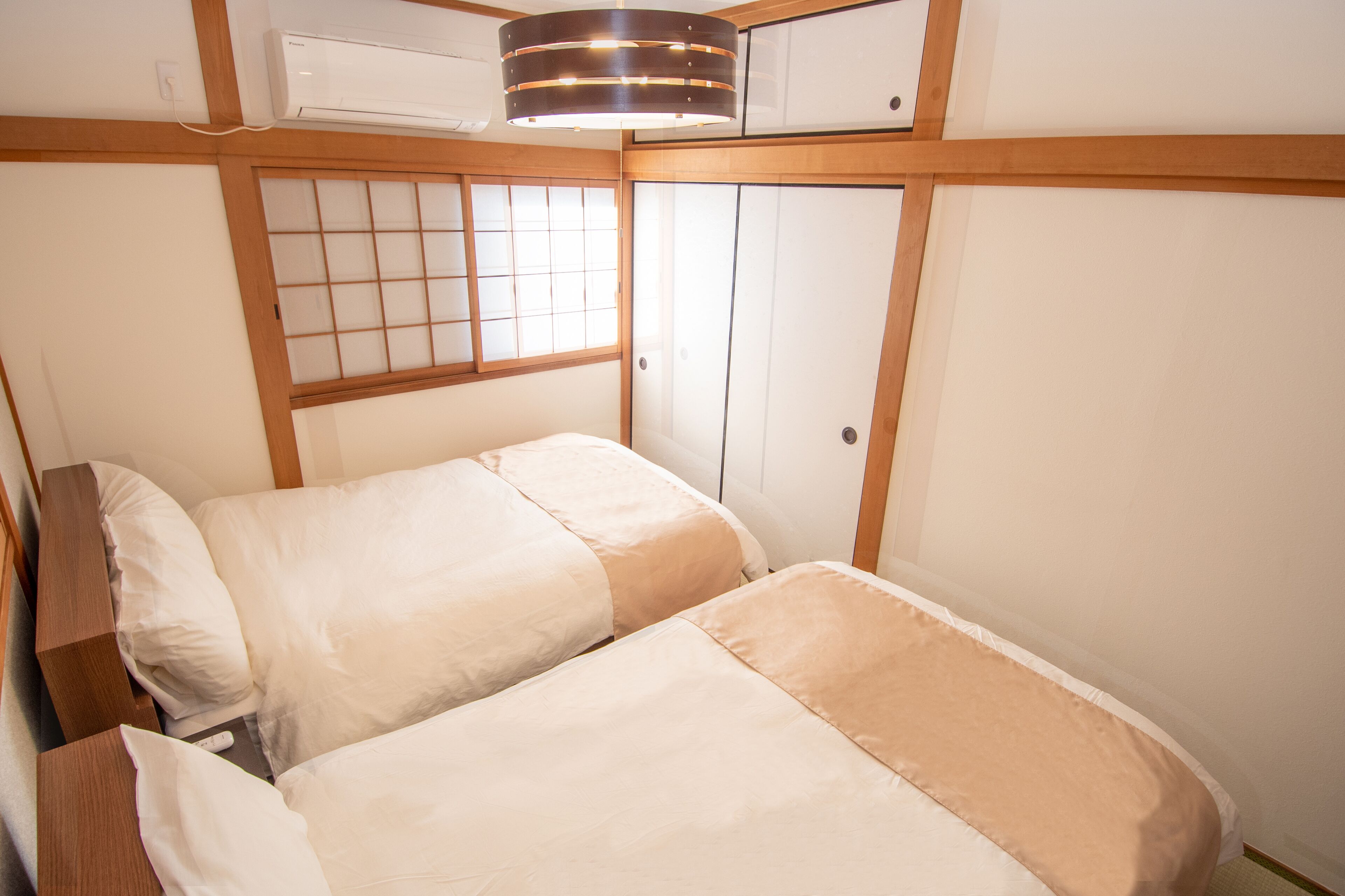 A room with two beds, a window with wooden frames, and a ceiling-mounted air conditioner.