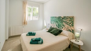 Ein Schlafzimmer mit Bett, einem Fenster mit Vorhängen, einem Heizkörper, einem Nachttisch mit Lampe und einer Wand mit einem großen Pflanzenbild.