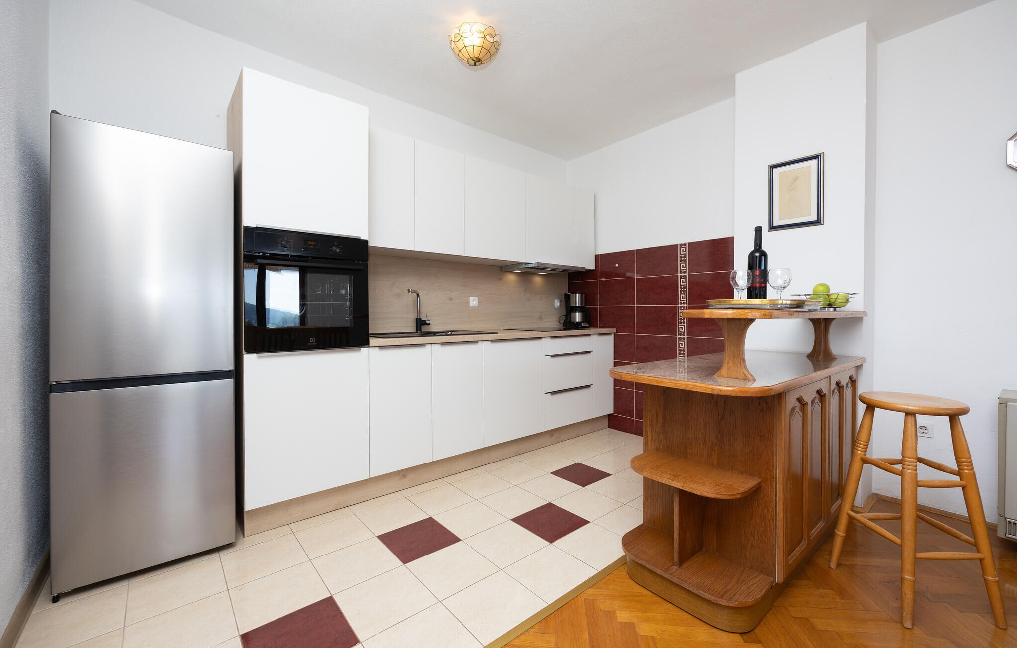 A modern kitchen with white cabinets, a stainless steel refrigerator, a built-in oven, a wooden bar counter, and a wooden stool.