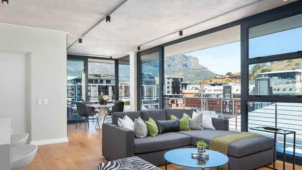 A modern living room with a grey sectional sofa, a round coffee table, and a zebra-patterned ottoman. Large windows offer a view of the city and mountains. - Docklands 502 (Cape Town)