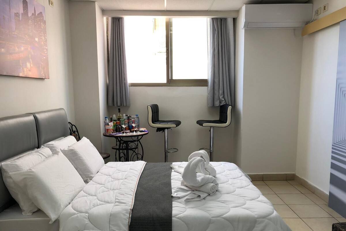 Quarto de hotel com cama, uma mesinha com artigos de higiene, duas cadeiras e uma janela com cortinas.