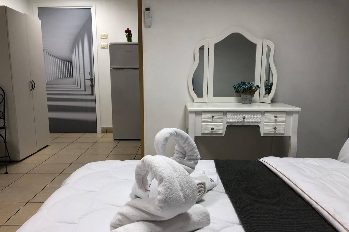 Quarto de hotel com uma cama, cômoda branca com espelho e um corredor que leva a outro quarto.