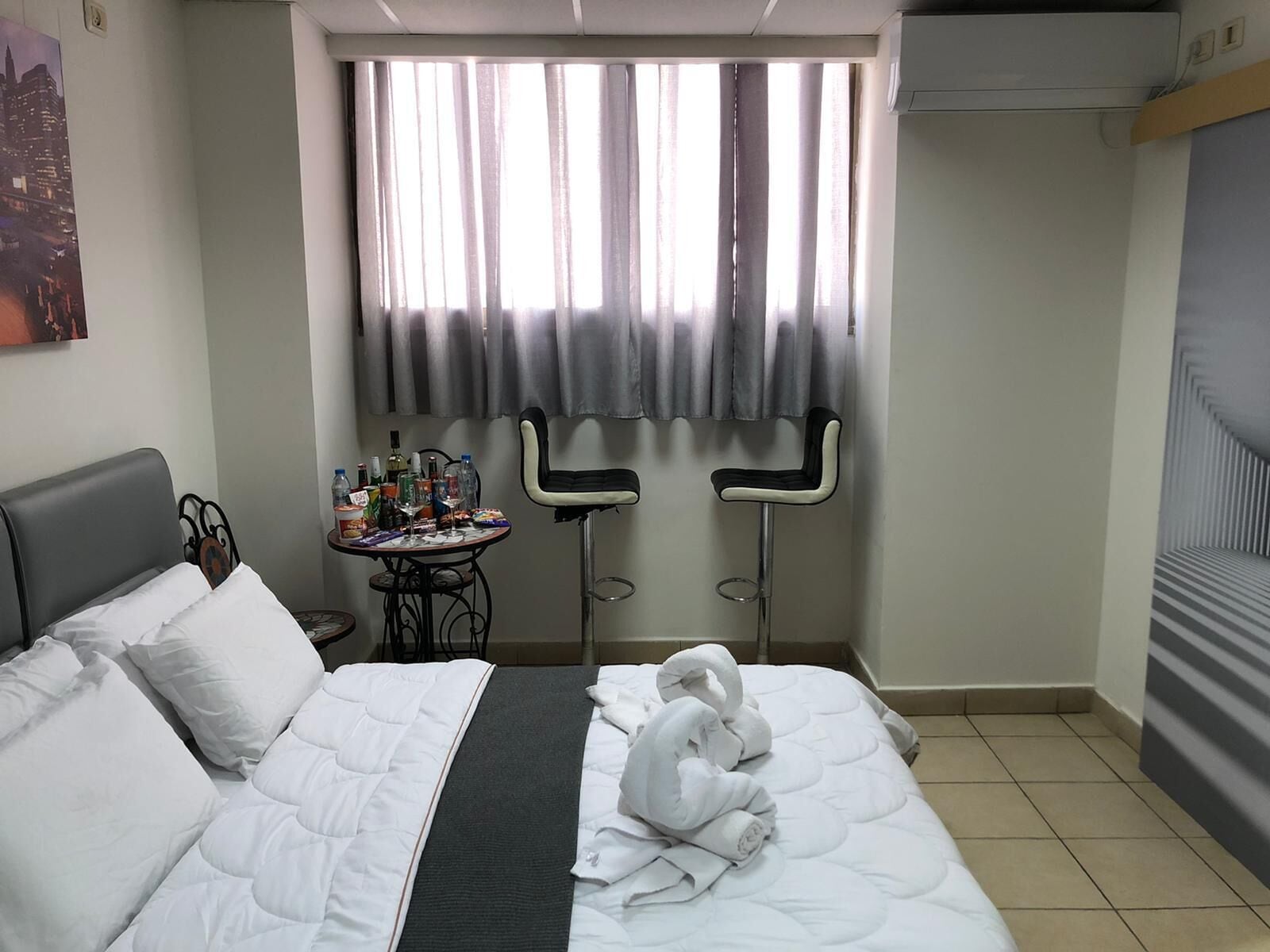 A hotel room with a bed, a small table with drinks, two bar stools, and a window with curtains.