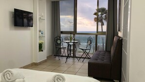 Een hotelkamer met een bed, een sofa, een televisie, een balkon met uitzicht op de oceaan, en een kleine tafel met stoelen.