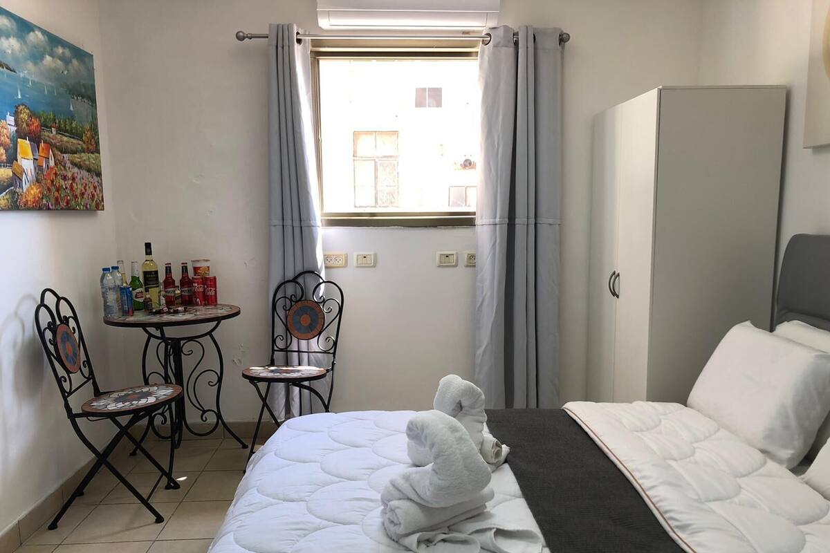 Quarto de hotel com cama, uma pequena mesa com cadeiras, um quadro na parede e uma janela com cortinas.