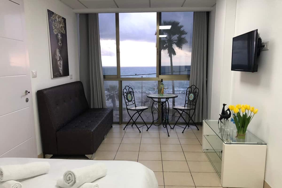 Quarto de hotel com cama, sofá, pequena mesa com cadeiras, televisão e vista para o mar.