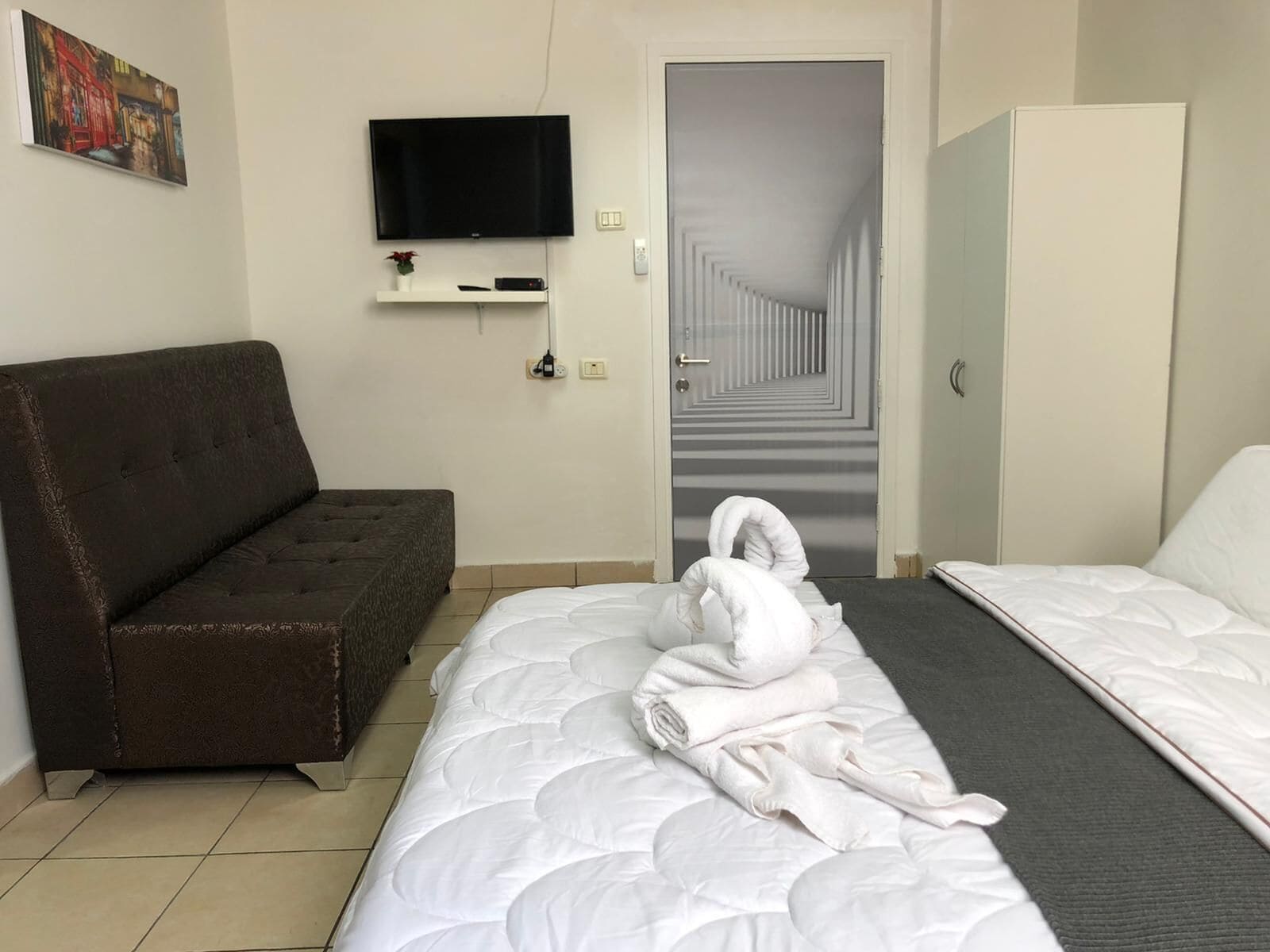 Hotellihuone, jossa on sänky, sohva, seinälle asennettu televisio ja ovi, joka johtaa käytävään.