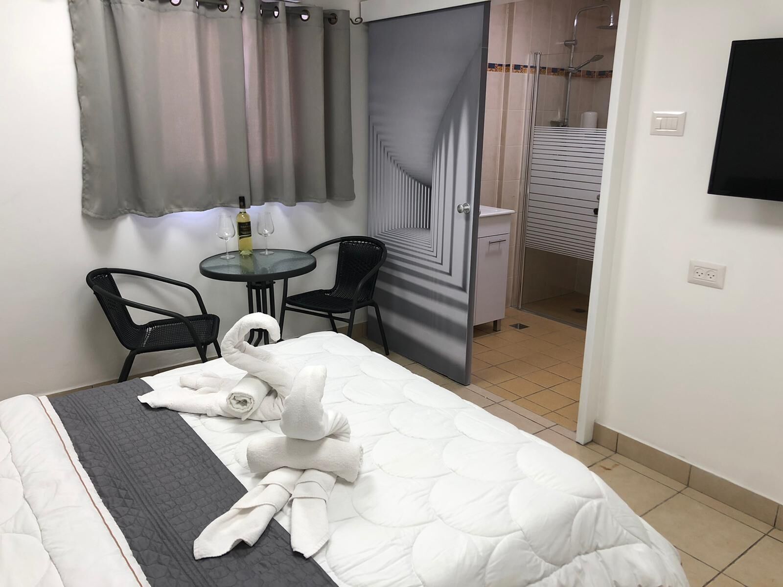 Habitación de hotel con una cama, dos sillas, una mesita con una botella y vasos, y un televisor en la pared.