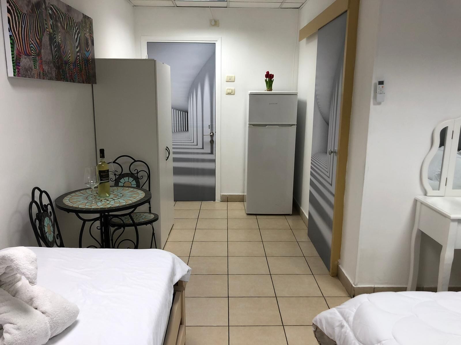 A hotel room with two beds, a small table with a bottle and glass, a refrigerator, and a painting of zebras on the wall.