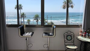 Een kamer met twee barkrukken en een kleine tafel met flessen, met uitzicht op een strand met palmbomen en de oceaan.