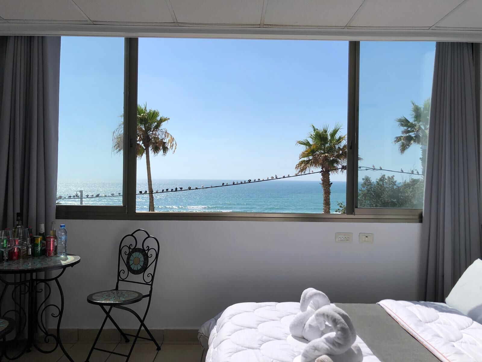 A hotel room with a large window overlooking the sea, a bed with a white duvet, a small table with drinks, and a chair.