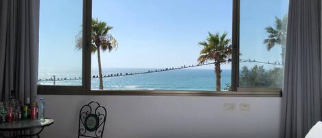 A hotel room with a large window overlooking the sea, a bed with a white duvet, a small table with drinks, and a chair.