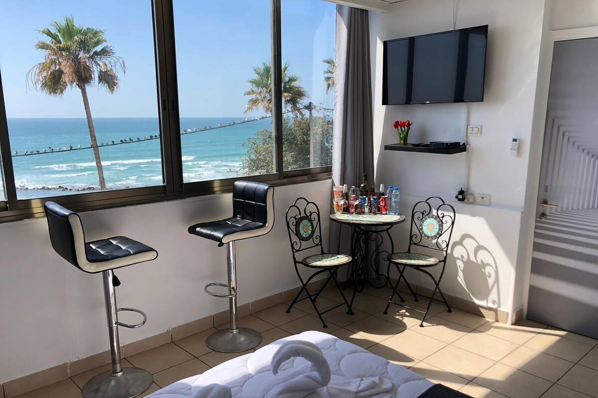 Quarto de hotel com uma cama, dois banquetes, uma mesinha com bebidas, uma televisão e vista para o oceano.