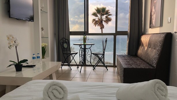 Een hotelkamer met een bed, een bank, een televisie, een tafel met stoelen, een raam met uitzicht op de zee en een palmboom, plus een potorkidee.