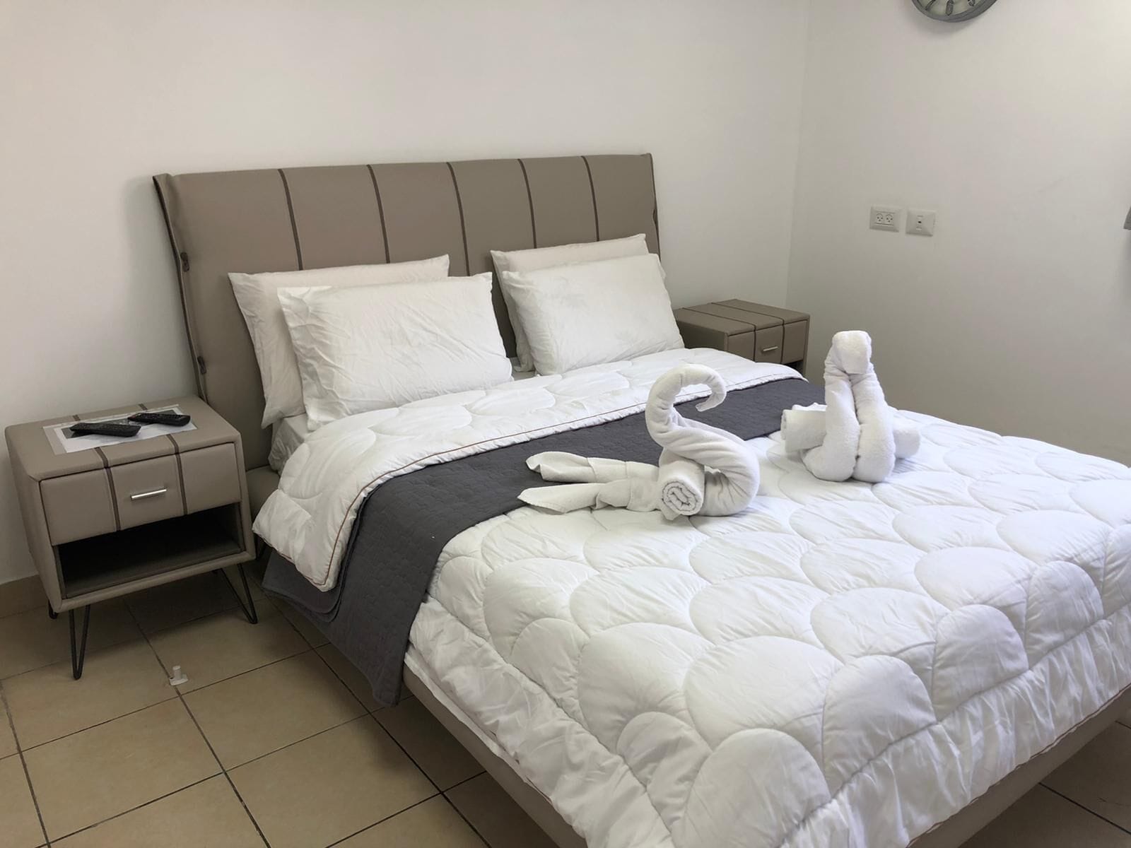 Un lit bien fait, avec une couette blanche matelassée, une tête de lit grise et deux oreillers blancs. On y trouve une petite table de chevet avec une télécommande et une serviette pliée.