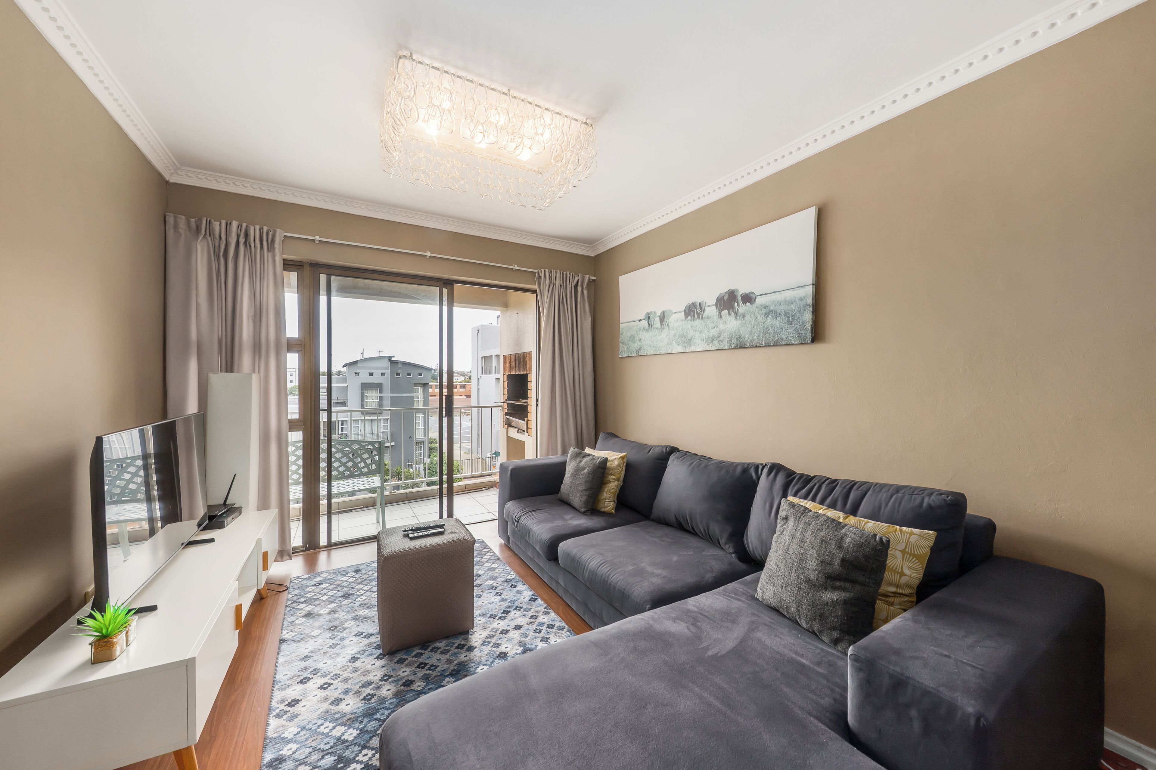 Ein modernes Wohnzimmer mit einem grauen Sofa, einem Flachbildfernseher, einem kleinen Tisch mit einer Pflanze und einem großen Fenster mit Vorhängen.