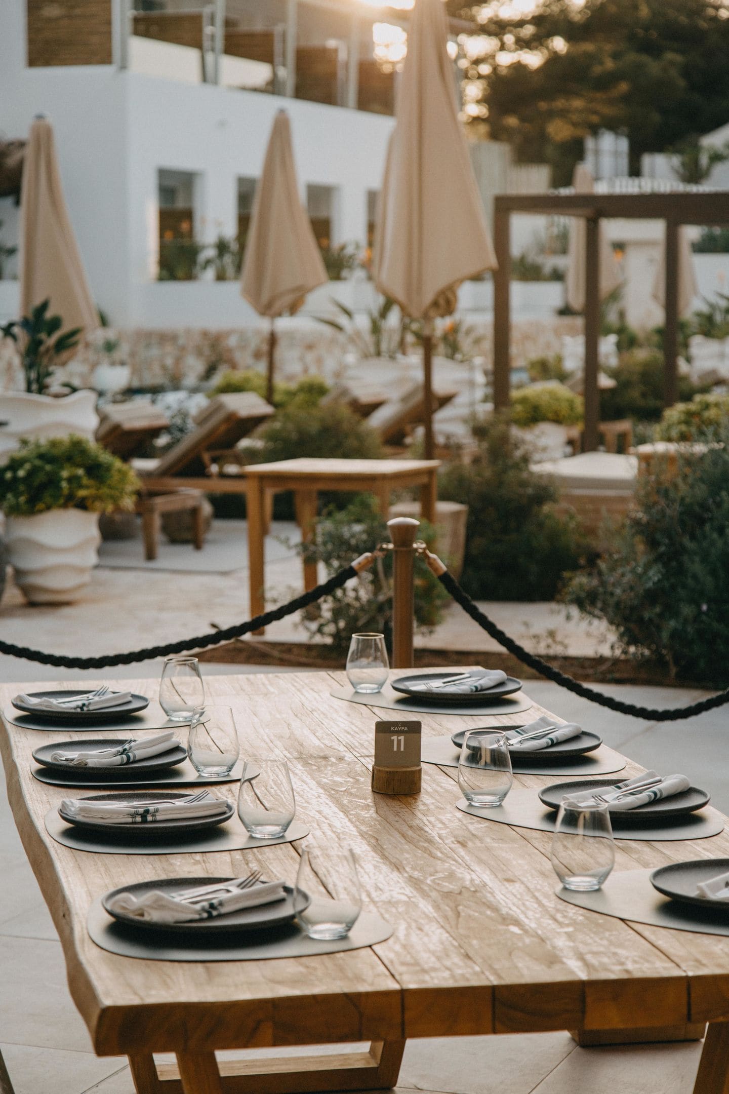 Mesa de comedor de madera dispuesta con platos, cubiertos y vasos, con el número 11, en un área de comedor al aire libre con plantas en macetas y sombrillas.