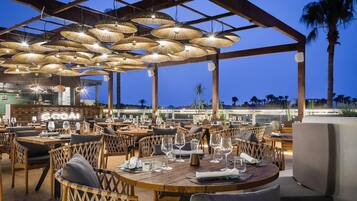 Un restaurante al aire libre con mesas redondas y sillas de mimbre, bajo una zona techada con lámparas colgantes.