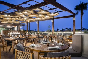 Un restaurante al aire libre con mesas redondas y sillas de mimbre, bajo una zona techada con lámparas colgantes.