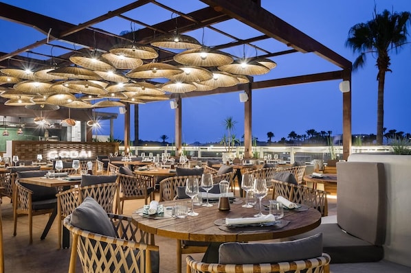 An outdoor restaurant with round tables and wicker chairs, under a covered area with hanging lamps. - Lago Resort Menorca Suites del Lago - Adults Only (Ciutadella de Menorca)