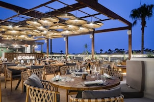 Un restaurante al aire libre con mesas redondas y sillas de mimbre, bajo una zona techada con lámparas colgantes.