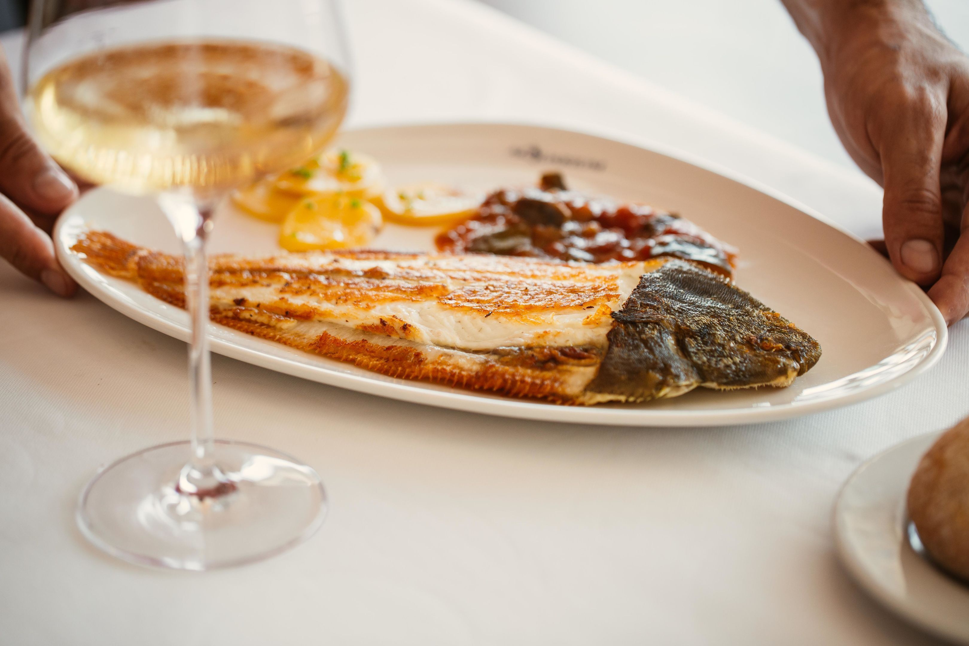 A plate of food with a fish, a glass of white wine, and a bread roll.