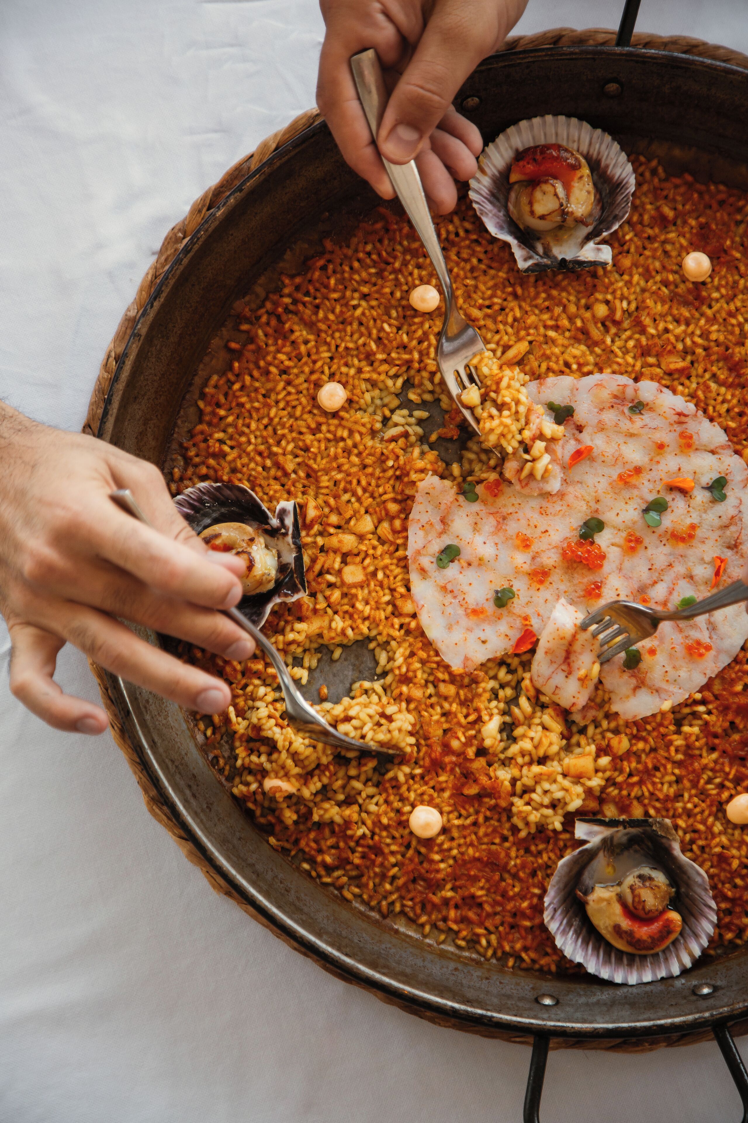 A large, round, metal pan filled with a rice-based dish, possibly paella, with several scallops and garnishes.