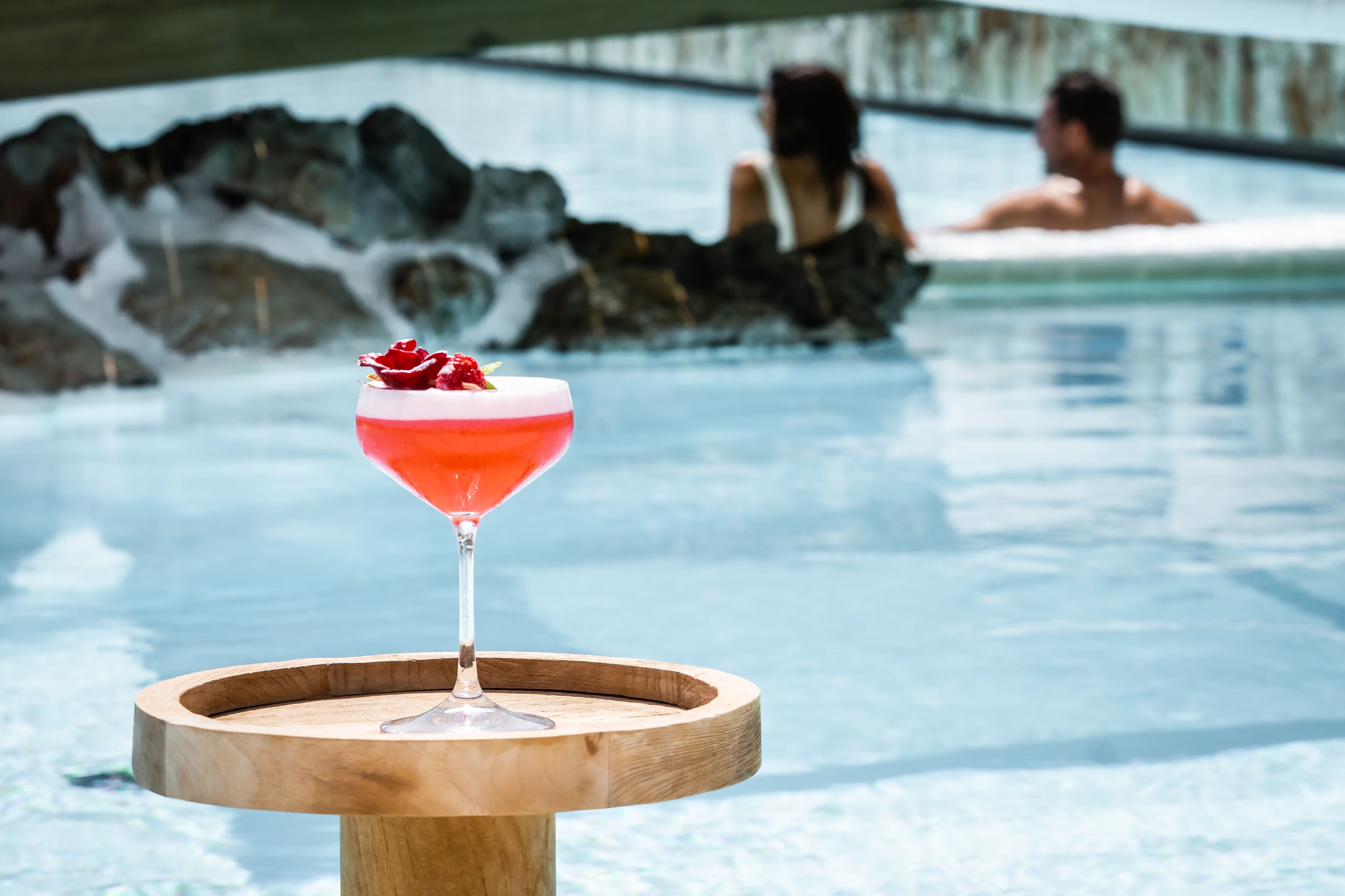 A cocktail on a wooden tray by a pool with a stone wall and a person in the background.