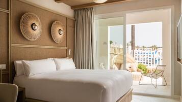 Habitación de hotel con una cama grande, dos lámparas circulares fijadas a la pared, un ventilador de techo y vistas a una playa y edificios de un complejo turístico a través de una puerta corrediza de vidrio.