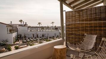 A rooftop terrace with a wicker chair and a wooden stool, overlooking a modern building complex with palm trees.