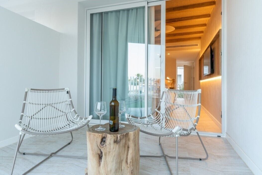 A modern room with two white wireframe chairs, a wooden stump table, and a bottle of wine.