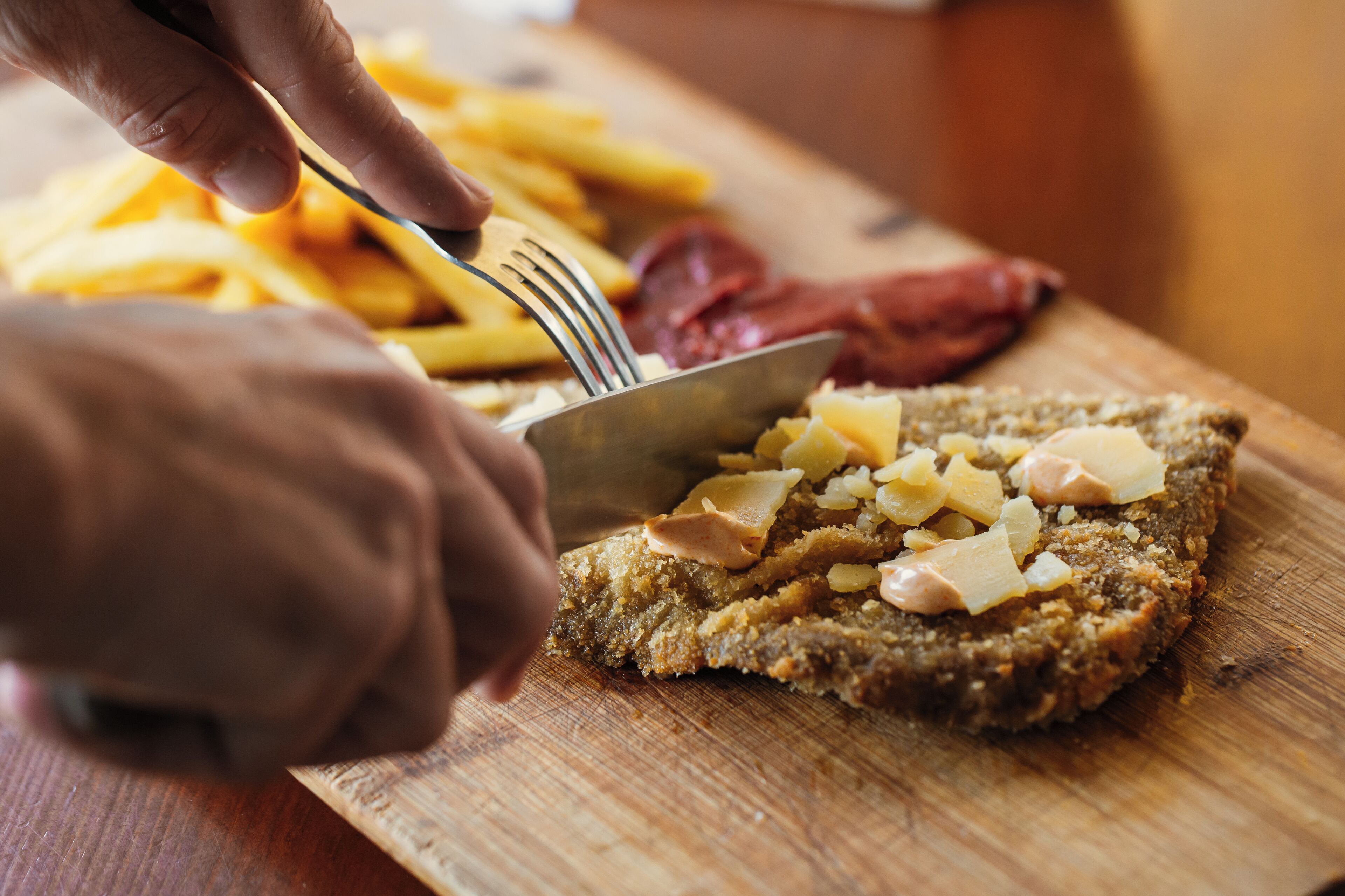 A wooden cutting board with a slice of bread topped with cheese and ham, and a hand using a fork to cut the bread.