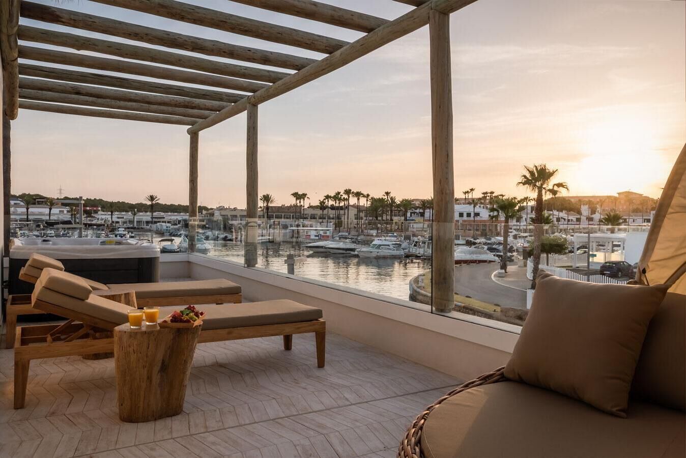 A balcony with wooden furniture, a view of a marina, and a sunset.