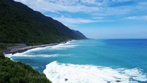 A coastal landscape with a rocky shoreline, a steep green hillside, and a clear blue sky.