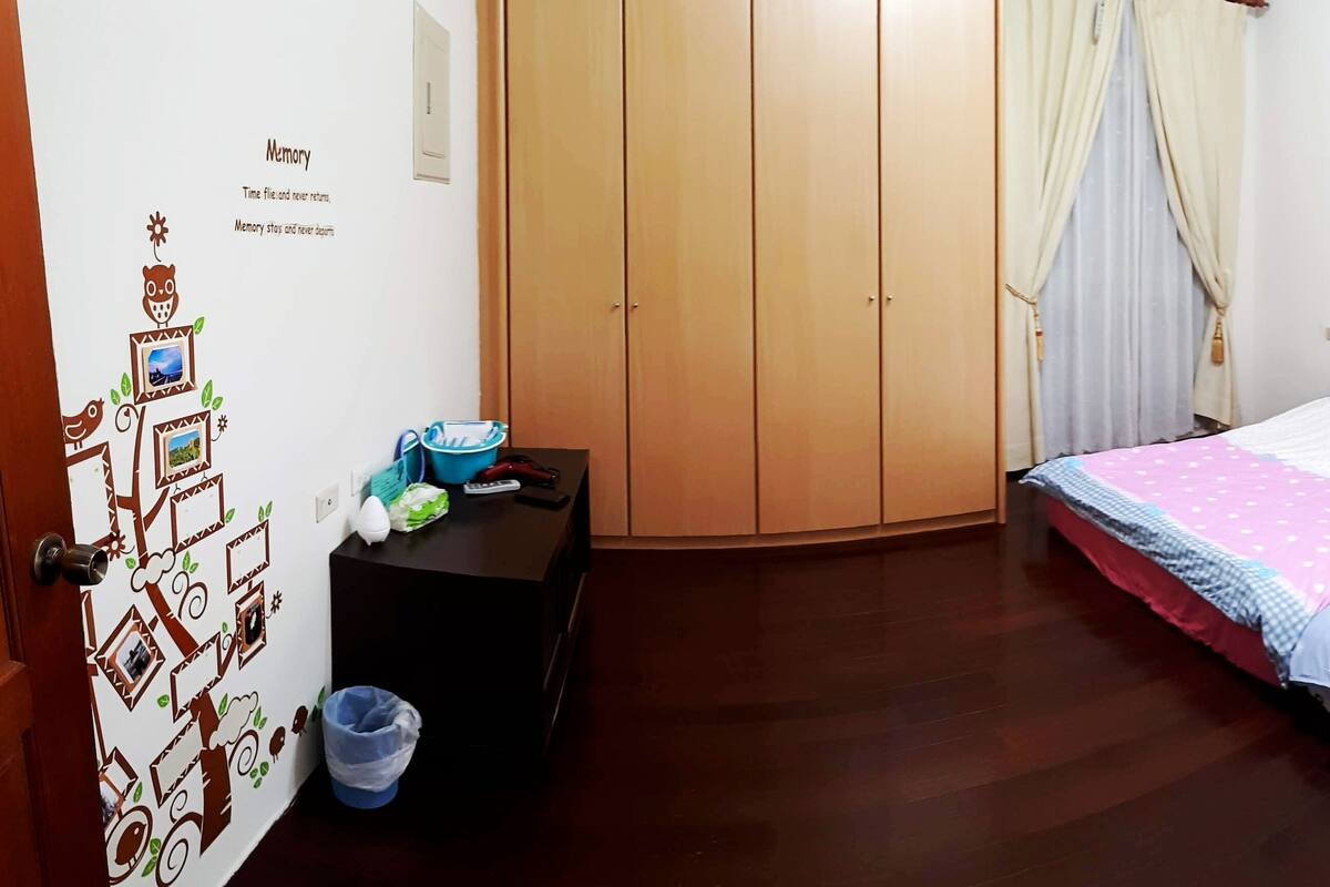Een slaapkamer met een bed, een kledingkast, een klein tafeltje met een blauwe emmer, en een muur met een boom en tekst.