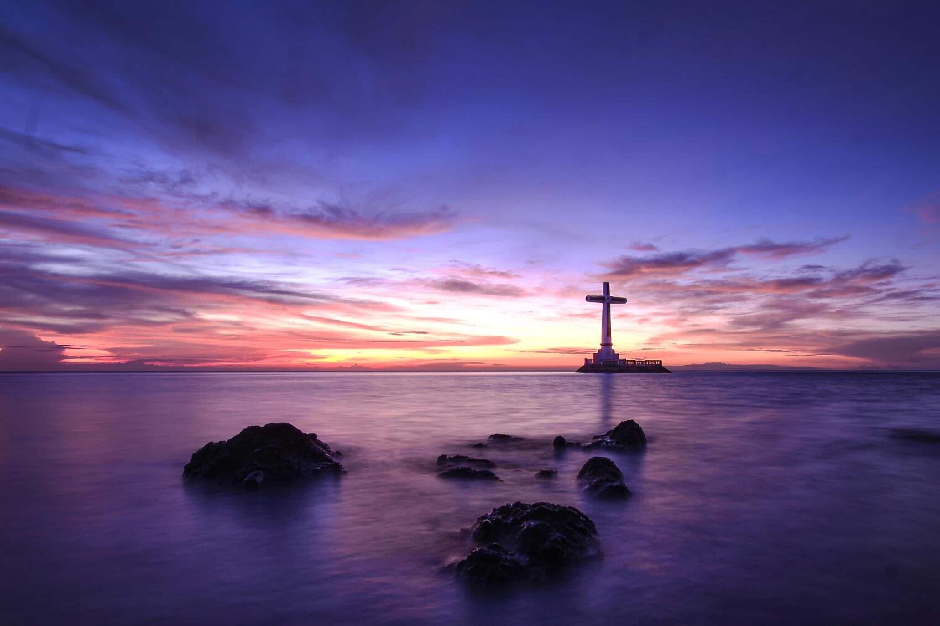A lighthouse at sunset with calm water and rocks in the foreground.